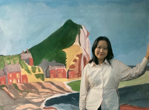 Woman in white shirt holding up green fabric in front of a colorful mural depicting a mountain village scene