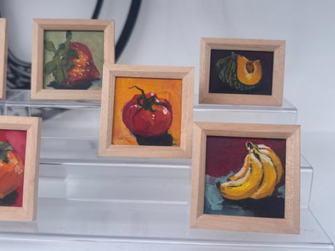 Collection of framed miniature paintings featuring red tomato, yellow bananas, and other produce displayed on white shelving